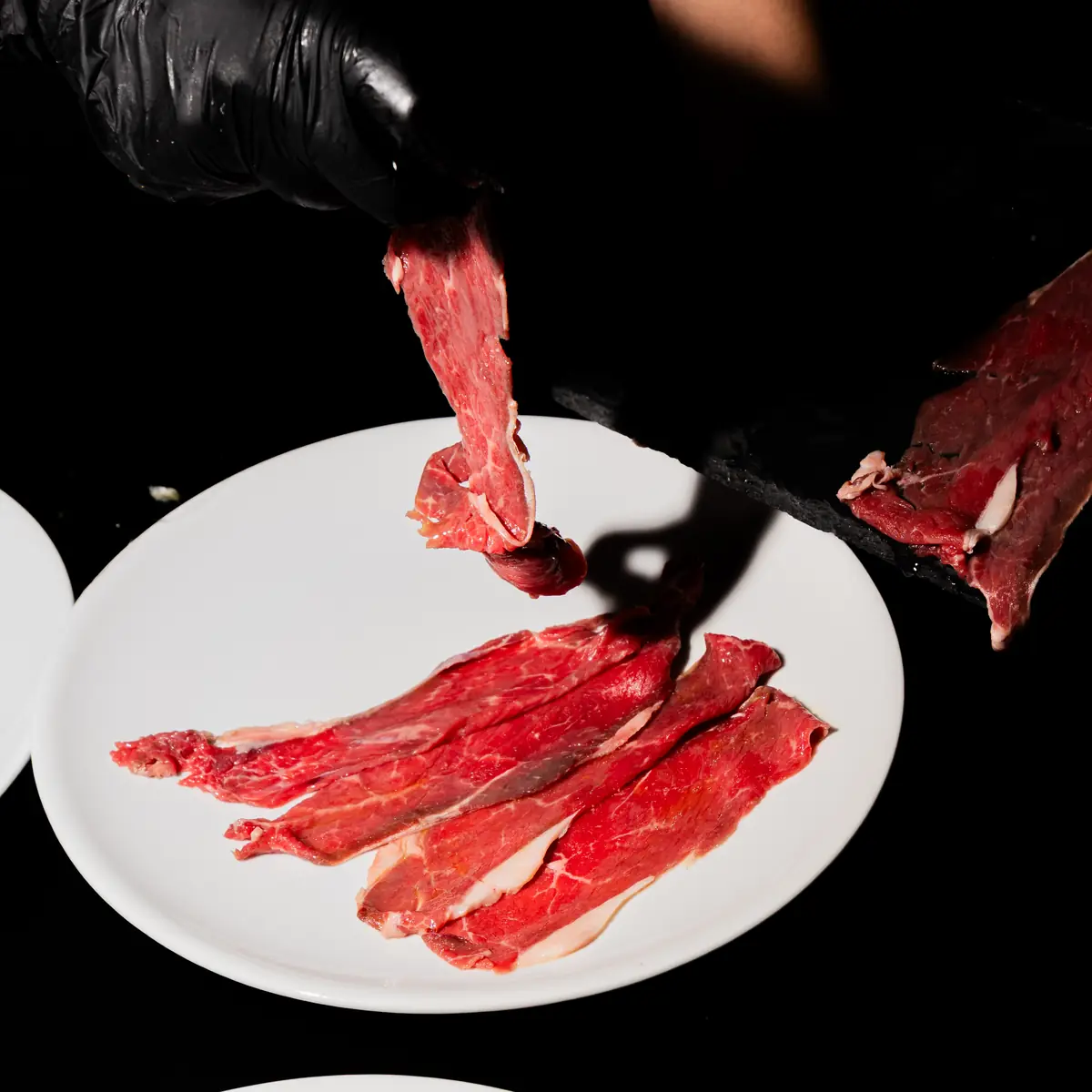 Una mano guantata posa fette di carne cruda su un piatto bianco
