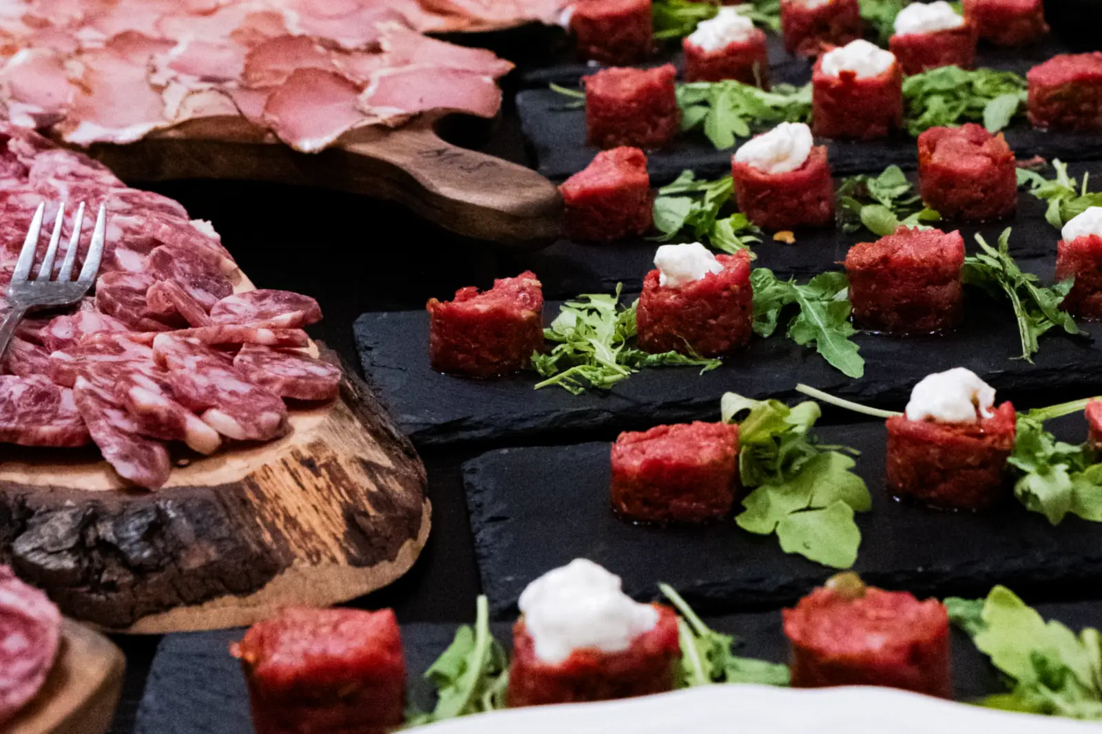 Tartare di carne a cubetti su lavagne nere, taglieri di salumi affettati e formaggi sulla tavola, su fondo nero